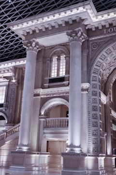 A grand architectural interior featuring large classical columns and intricately detailed arches made of stone. The structure includes ornate designs and a high ceiling with a geometric pattern. Soft lighting highlights the elaborate carvings and intricate moldings on the pillars and arches.