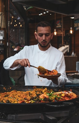 A chef wearing a white uniform is serving a dish from a large pan filled with a colorful and hearty paella. The dish is rich with ingredients such as prawns, mussels, and vegetables, creating a vibrant and appetizing display. The background suggests a busy kitchen or food market environment.
