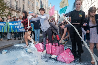 A group of people are participating in a protest, pouring milk onto the street. They are surrounded by banners and flags. The setting appears to be a city street lined with buildings.