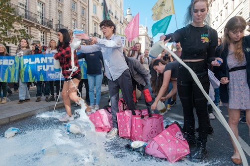 A group of people are participating in a protest, pouring milk onto the street. They are surrounded by banners and flags. The setting appears to be a city street lined with buildings.