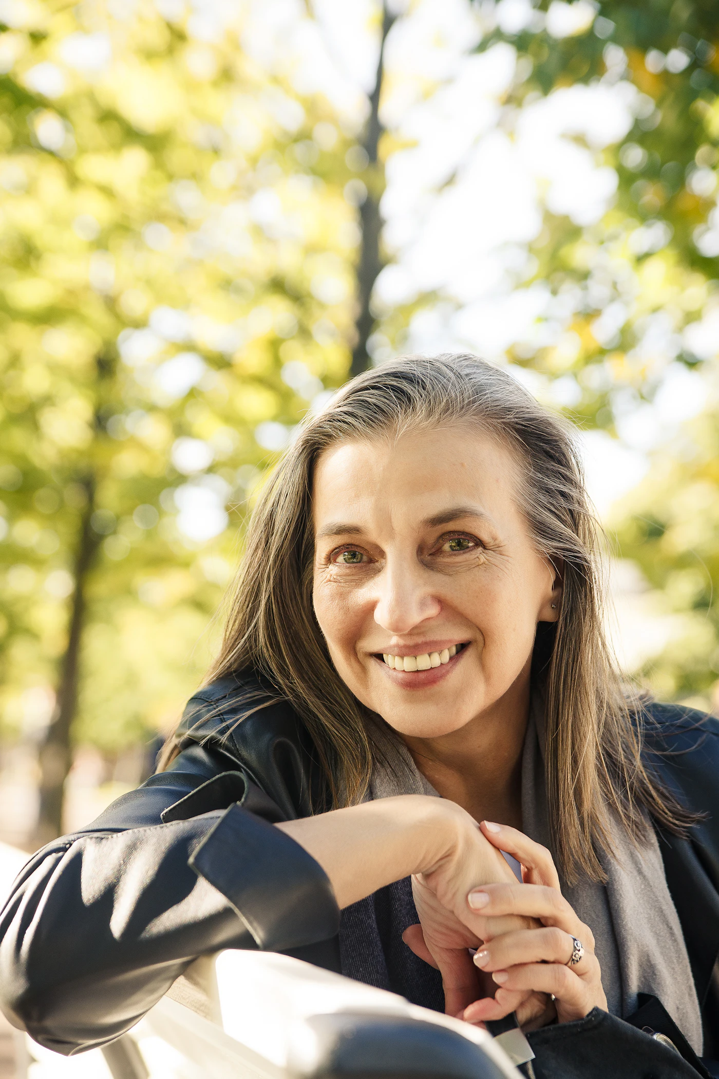 A woman of about sixty with long grey hair, smiling broadly among sunlit trees
