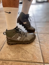 A person's legs wearing white socks and gray high-top athletic shoes standing on a tiled floor. The shoes and socks have distinct brand logos.