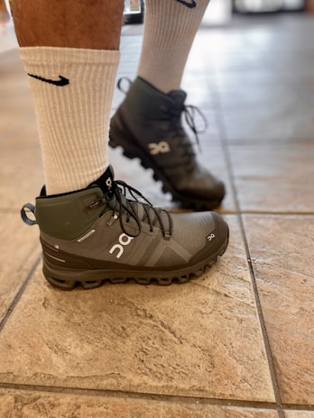 A person's legs wearing white socks and gray high-top athletic shoes standing on a tiled floor. The shoes and socks have distinct brand logos.