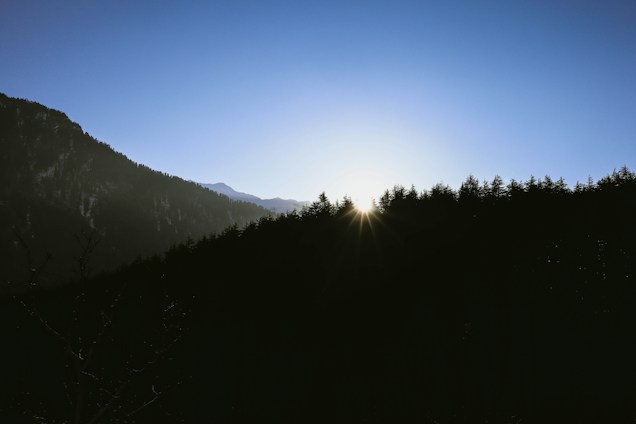A tranquil mountain scene at sunrise with the sun peeking over the horizon, casting silhouettes of trees and hills. The sky is a clear blue, and the light creates a serene and peaceful atmosphere.