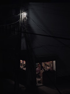 A dimly lit scene featuring a building with numerous dark wires crossing the frame. Part of the building is illuminated by a single bright light up high, and below, there is a small shop or room visible through a doorway. The interior is faintly visible, with shelves stocked with various items and two chairs visible inside.