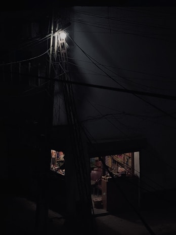 A dimly lit scene featuring a building with numerous dark wires crossing the frame. Part of the building is illuminated by a single bright light up high, and below, there is a small shop or room visible through a doorway. The interior is faintly visible, with shelves stocked with various items and two chairs visible inside.