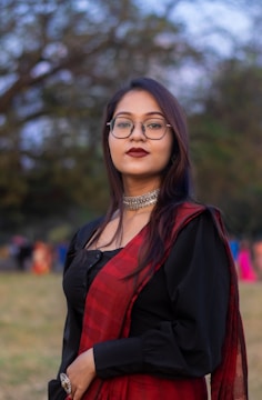 A woman wearing glasses, a black blouse with long sleeves, and a red saree is standing outdoors. She has long dark hair and is wearing dark lipstick and a silver choker necklace. The background is slightly blurred, showing trees and other people in the distance.