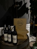 Wine bottles are placed on a wooden barrel next to stacked pizza boxes with the label 'Pizza Locale.' The setting includes a dimly lit ambiance with a potted plant nearby and a tiled wall in the background.