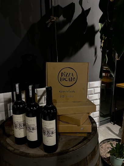 Wine bottles are placed on a wooden barrel next to stacked pizza boxes with the label 'Pizza Locale.' The setting includes a dimly lit ambiance with a potted plant nearby and a tiled wall in the background.