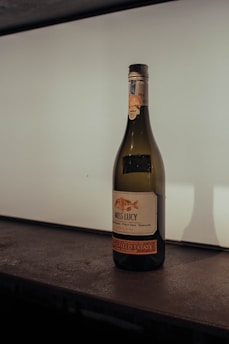 A wine bottle labeled 'Miss Lucy' is placed on a dark, textured surface. The bottle has a fish illustration on the label and contains details about the wine's blend, including Pinot Gris and Semillon. The background is softly lit with a gradient, providing subtle contrast to the wine bottle's green glass and detailed label.