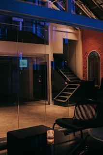 An interior setting featuring a modern, minimalistic design with an industrial feel. A metal staircase ascends into darkness, and there is a black chair with a ribbed backrest in the foreground. A candle on a glossy table adds a soft, warm glow. The walls are a mix of different textures, including glass and a red, textured surface.