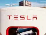 A Tesla charging station is prominently displayed with the logo clearly visible in bold red letters. The background features a blue sky and a blurry sign, while the charging cable is partially visible at the bottom of the charging unit.