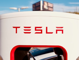 A Tesla charging station is prominently displayed with the logo clearly visible in bold red letters. The background features a blue sky and a blurry sign, while the charging cable is partially visible at the bottom of the charging unit.