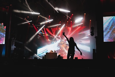 A stage with vibrant lighting effects, featuring a large, colorful digital screen displaying abstract patterns. A DJ setup is in the center, with a silhouette of a person dancing energetically in the foreground. Lights of various colors, including red and white, create a dynamic atmosphere.