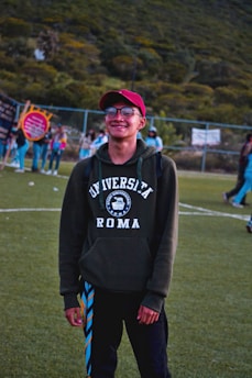 A person is standing on a grassy field wearing a red cap and a dark green hoodie with 'Universita Roma' printed on it. There is a group of people in the background, suggesting a social or recreational setting. The scene is outdoors, with a hilly, green landscape behind a chain-link fence.