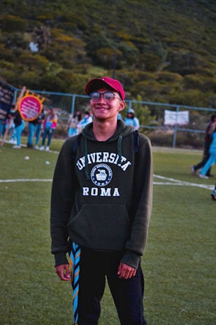 A person is standing on a grassy field wearing a red cap and a dark green hoodie with 'Universita Roma' printed on it. There is a group of people in the background, suggesting a social or recreational setting. The scene is outdoors, with a hilly, green landscape behind a chain-link fence.