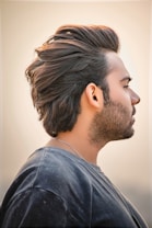 A man with dark brown, voluminous hair is captured in a side profile. The hairstyle is styled back with noticeable texture. He has a beard and is wearing a casual, dark-colored T-shirt. The background is softly blurred, giving a warm, neutral tone.
