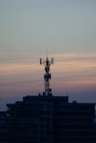 A telecommunications tower is perched on top of a high-rise building against a backdrop of a dusky sky. The sky is painted with gradients of blue and orange, reflecting the transition from day to night.