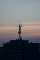 A telecommunications tower is perched on top of a high-rise building against a backdrop of a dusky sky. The sky is painted with gradients of blue and orange, reflecting the transition from day to night.