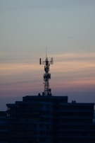 A telecommunications tower is perched on top of a high-rise building against a backdrop of a dusky sky. The sky is painted with gradients of blue and orange, reflecting the transition from day to night.