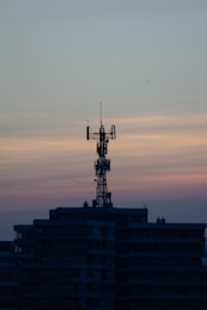 A telecommunications tower is perched on top of a high-rise building against a backdrop of a dusky sky. The sky is painted with gradients of blue and orange, reflecting the transition from day to night.