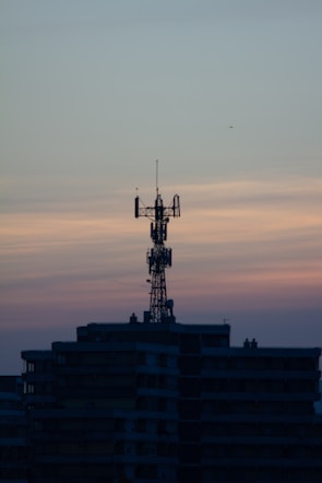 A telecommunications tower is perched on top of a high-rise building against a backdrop of a dusky sky. The sky is painted with gradients of blue and orange, reflecting the transition from day to night.
