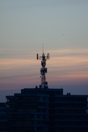 A telecommunications tower is perched on top of a high-rise building against a backdrop of a dusky sky. The sky is painted with gradients of blue and orange, reflecting the transition from day to night.