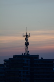 A telecommunications tower is perched on top of a high-rise building against a backdrop of a dusky sky. The sky is painted with gradients of blue and orange, reflecting the transition from day to night.