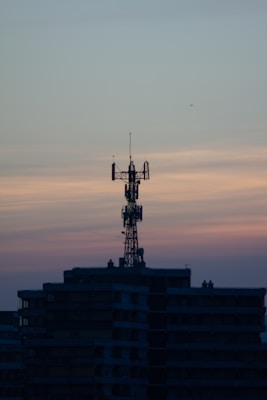 A telecommunications tower is perched on top of a high-rise building against a backdrop of a dusky sky. The sky is painted with gradients of blue and orange, reflecting the transition from day to night.
