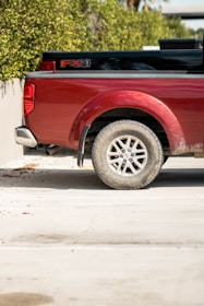 A red pickup truck is parked next to a green hedge, with only the rear part of the vehicle visible. The truck has a logo on the side and features a dusty tire. The background includes a concrete wall and the sunlight is casting shadows on the ground.
