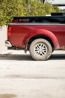 A red pickup truck is parked next to a green hedge, with only the rear part of the vehicle visible. The truck has a logo on the side and features a dusty tire. The background includes a concrete wall and the sunlight is casting shadows on the ground.