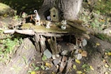 A small, whimsical fairy house is crafted at the base of a tree trunk, surrounded by natural elements. It features wooden and mossy components, with tiny decorative birdhouses and miniature figurines placed on top. The setting is outdoors, nestled within a forested area with scattered leaves and dappled sunlight.
