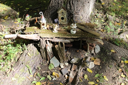 A small, whimsical fairy house is crafted at the base of a tree trunk, surrounded by natural elements. It features wooden and mossy components, with tiny decorative birdhouses and miniature figurines placed on top. The setting is outdoors, nestled within a forested area with scattered leaves and dappled sunlight.