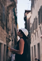 A person wearing a white hat and a black outfit is standing between two tall, narrow buildings. They are holding a small book or pamphlet and appear to be looking upwards, possibly observing the architecture or surroundings. The scene is sunlit, with a clear blue sky visible above.
