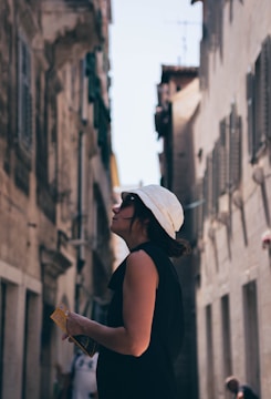 A person wearing a white hat and a black outfit is standing between two tall, narrow buildings. They are holding a small book or pamphlet and appear to be looking upwards, possibly observing the architecture or surroundings. The scene is sunlit, with a clear blue sky visible above.
