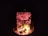 A campfire burning brightly at night, illuminating a foil-wrapped object positioned directly over the flames. The fire emits a warm glow with visible flames and embers against the dark background.