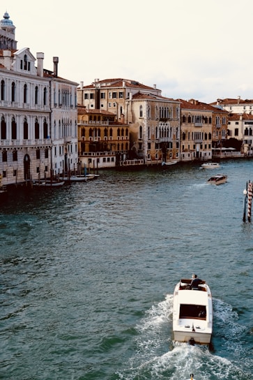 A canal is lined with historical buildings featuring classical architecture, displaying arched windows and ornate details. A motorboat is moving along the water, creating a small wake. The scene is serene, capturing the essence of a charming, old-world city reflected in the soft light.