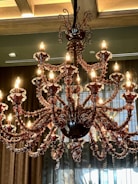 An ornate chandelier with multiple lit bulbs displayed in a luxurious setting. It features intricate glass arms and decorative elements in a dark, rich color. The background includes draped curtains and a ceiling with wooden beams.