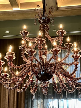 An ornate chandelier with multiple lit bulbs displayed in a luxurious setting. It features intricate glass arms and decorative elements in a dark, rich color. The background includes draped curtains and a ceiling with wooden beams.