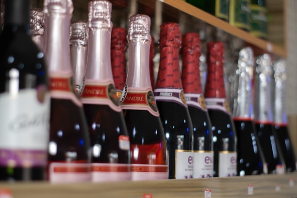 A shelf is stocked with various bottles of sparkling wine or champagne, featuring different labels and foil wrappings. The bottles are closely arranged in a neat row, suggesting a selection of sparkling beverages.