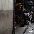 A dimly lit alleyway featuring people engaged in various activities. One person is bending down beside a motorcycle, while others are sitting or resting. Bright yellow and green accents from umbrellas or covers contrast with the otherwise dark environment. The scene captures a sense of everyday street life in an urban setting.