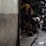 A dimly lit alleyway featuring people engaged in various activities. One person is bending down beside a motorcycle, while others are sitting or resting. Bright yellow and green accents from umbrellas or covers contrast with the otherwise dark environment. The scene captures a sense of everyday street life in an urban setting.