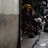 A dimly lit alleyway featuring people engaged in various activities. One person is bending down beside a motorcycle, while others are sitting or resting. Bright yellow and green accents from umbrellas or covers contrast with the otherwise dark environment. The scene captures a sense of everyday street life in an urban setting.