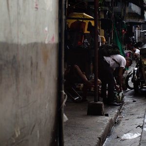 A dimly lit alleyway featuring people engaged in various activities. One person is bending down beside a motorcycle, while others are sitting or resting. Bright yellow and green accents from umbrellas or covers contrast with the otherwise dark environment. The scene captures a sense of everyday street life in an urban setting.