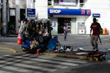A street vendor stall is set up on a sidewalk, displaying various goods like bags, necklaces, and other accessories. Two people are nearby, one sitting on the ground sorting items and another walking past carrying an umbrella. In the background, there's a shop entrance with signs for services including a restroom.
