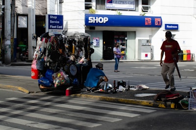 A street vendor stall is set up on a sidewalk, displaying various goods like bags, necklaces, and other accessories. Two people are nearby, one sitting on the ground sorting items and another walking past carrying an umbrella. In the background, there's a shop entrance with signs for services including a restroom.