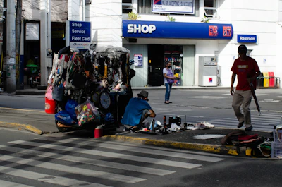A street vendor stall is set up on a sidewalk, displaying various goods like bags, necklaces, and other accessories. Two people are nearby, one sitting on the ground sorting items and another walking past carrying an umbrella. In the background, there's a shop entrance with signs for services including a restroom.