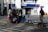 A street vendor stall is set up on a sidewalk, displaying various goods like bags, necklaces, and other accessories. Two people are nearby, one sitting on the ground sorting items and another walking past carrying an umbrella. In the background, there's a shop entrance with signs for services including a restroom.