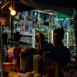A dimly lit market stall where a person is preparing a drink. Several blenders with ingredients are visible, and there are fruits like melons on the table. The background shows a display of sunglasses with various advertisements above.