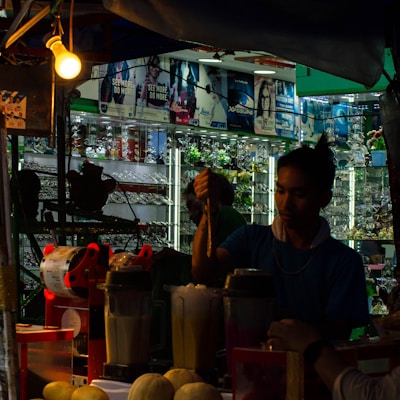 A dimly lit market stall where a person is preparing a drink. Several blenders with ingredients are visible, and there are fruits like melons on the table. The background shows a display of sunglasses with various advertisements above.